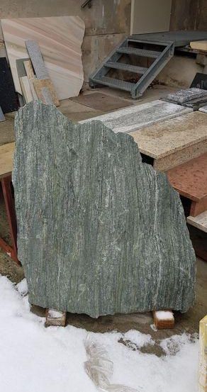 Ein großer, rauer grüner Steinblock liegt auf dem Boden mit Schnee. Dahinter befindet sich eine Treppe mit Metallhandläufen und einer Betonwand. In der Nähe sind verschiedene Materialien wie Holz, Metall und Betonblöcke.