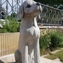 Eine Betonstatue eines Hundes sitzt auf einer Betonfläche in einem Garten, umgeben von Pflanzen und einem Metallzaun. Hinter dem Zaun ist ein Auto geparkt, und es gibt Bäume und ein Gebäude.