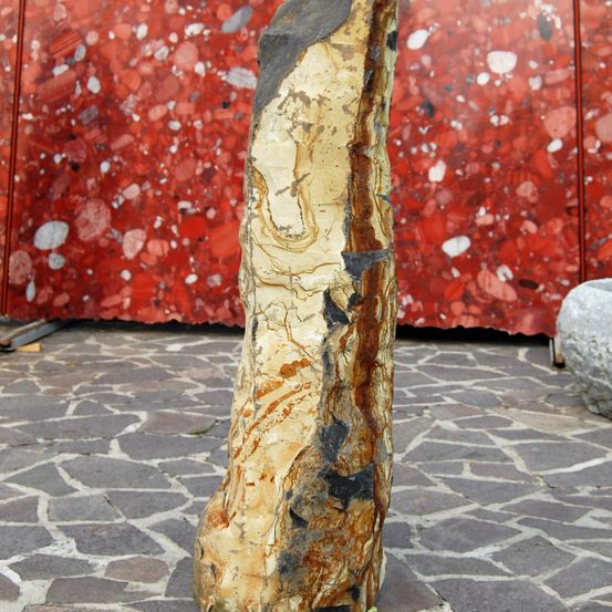 Ein großer, unregelmäßig geformter Stein mit rotbrauner und grauer Färbung steht auf einem Kopfsteinpflaster, mit einem roten Marmorhintergrund, der kleine weiße und schwarze Flecken aufweist.