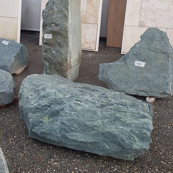 Verschiedene Steine sind im Freien auf einer Kiesoberfläche ausgestellt. Die Steine variieren in Form und Größe, einige sind groß und andere kleiner. Sie sind nummeriert, und im Hintergrund befindet sich ein Holzpfosten.