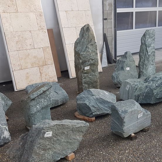 Verschiedene grüne Steinblöcke mit weißen Etiketten und Nummern sind auf dem Boden angeordnet. Dahinter befinden sich rechteckige Steinfliesen und eine Garage mit Glasfenstern.