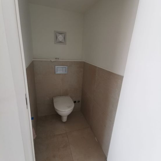 Ein leeres Badezimmer mit einer weißen Toilette und beigen Fliesen an Wänden und Boden. Über der Toilette befindet sich ein weißer Lüftungsschacht.