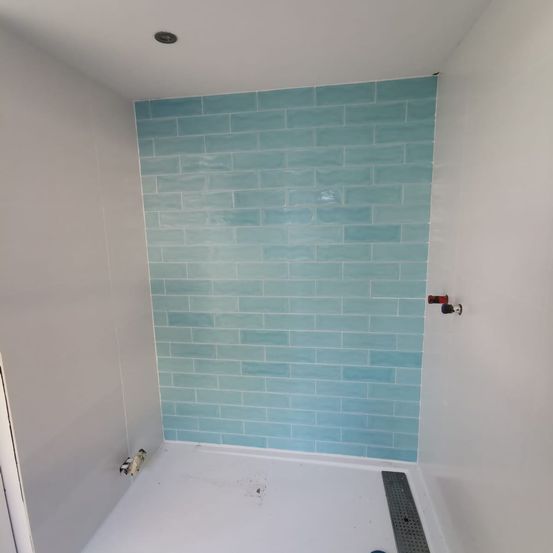 Ein Badezimmer mit weißen Wänden und einer weißen Duschbasis, ausgestattet mit einer türkisfarbenen gefliesten Wand. Ein Abfluss und ein Wasserhahn sind an der Wand sichtbar.