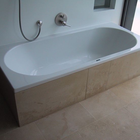 Eine weiße Badewanne mit Wasserhahn steht an einer Wand mit beigen Fliesen. Ein Duschkopf ist am Wasserhahn befestigt.
