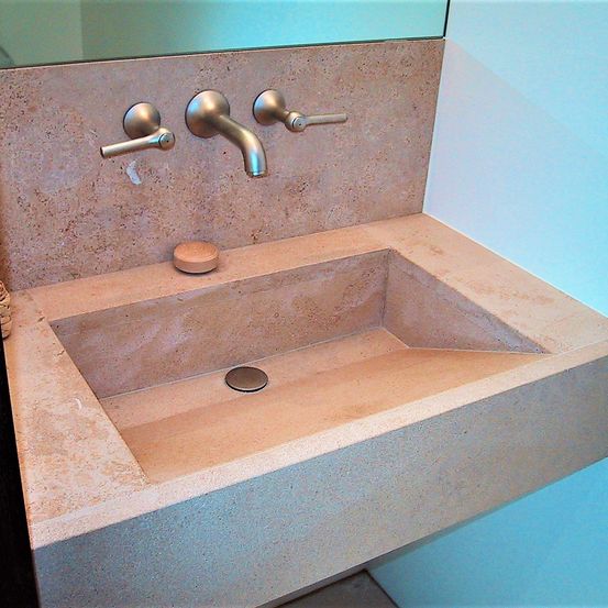 Ein modernes Badezimmer mit einem Betonwaschbecken und zwei silbernen Armaturen. Ein kleiner brauner Seifenspender und ein Abfluss befinden sich im Waschbecken. Die Wand ist blau gestrichen.