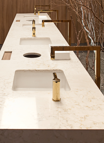Eine Reihe von vier weißen Waschbecken mit goldenen Armaturen auf einer Marmortheke. Hinter den Waschbecken ist ein Baumzweig zu sehen, und der Hintergrund besteht aus einer Holzwand.