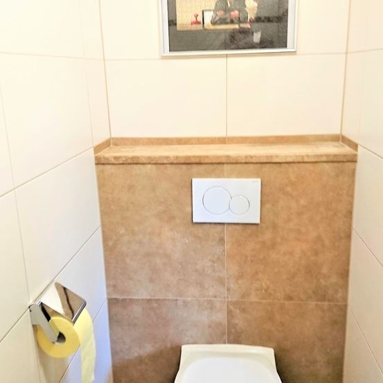 Ein kleines Badezimmer mit beigen Fliesen an Wand und Boden. Es gibt eine Toilettenschüssel und ein weißes Schalterpanel, das über der Toilette an der Wand montiert ist. Eine Rolle Toilettenpapier ist an der Wand montiert.