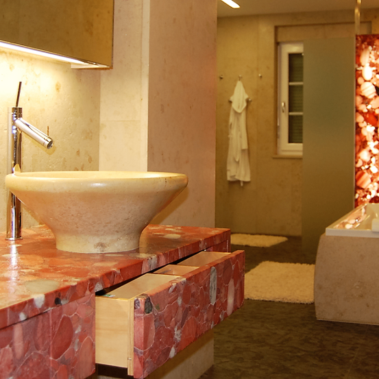 Ein modernes Badezimmer mit einem großen Becken auf einer roten Marmor-Arbeitsplatte. Ein Handtuch hängt neben einem Fenster mit Rolläden. Eine Badewanne und ein weiterer Teppich sind zur Seite.