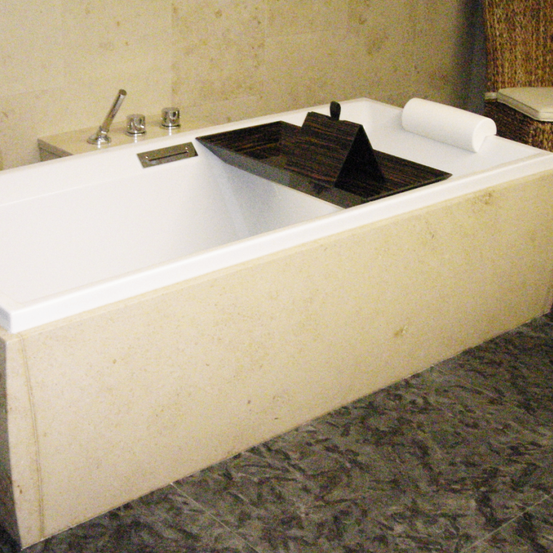Eine große weiße Badewanne mit einem Holztablett steht in einem Badezimmer mit gefliestem Boden und beigefarbenen Wänden.