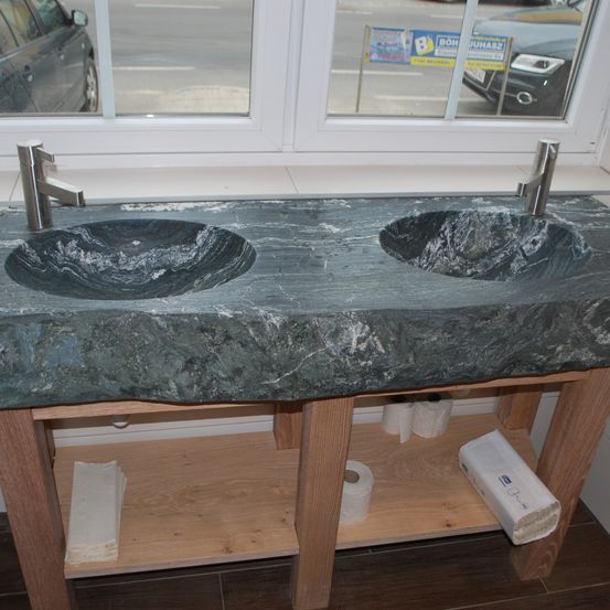 Ein Doppelbecken-Steinarbeitsplatte mit zwei Wasserhähnen. Darunter befindet sich ein Holzregal mit zwei Rollen Toilettenpapier und einer Packung Taschentücher. Das Waschbecken spiegelt das Bild zweier Autos auf der Straße wider.