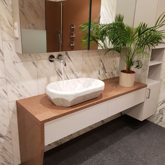Ein Badezimmer mit einem weißen Marmorwaschbecken, einem Topf mit einer Pflanze und einem Spiegel darüber. Das Waschbecken befindet sich über einem Holzarbeitsplatten mit Schränken darunter. Die Wände sind mit Marmorfliesen und einer Steckdose verkleidet.