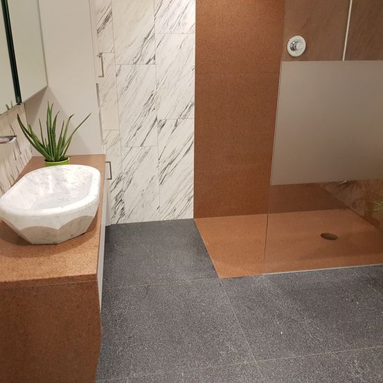 Ein Badezimmer mit einem weißen Marmorwaschbecken und einer grünen Pflanze auf einem braunen Counter. Die Wände sind marmorartig gemustert, und die Dusche ist mit Glas umschlossen.