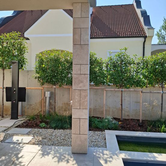 Ein Außenbereich mit einer Betonmauer, der gepflegte Bäume, ein kleiner Pool und ein Weg zum Haus umfasst.