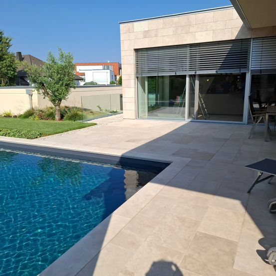 Ein modernes Haus mit einer Glastür und Fenstern blickt auf einen sauberen, blauen Swimmingpool. Fliesenböden umgeben den Pool. Bäume und ein Rasenbereich sind in der Nähe.