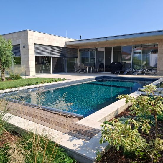 Ein modernes Haus mit Swimmingpool, Außenterrasse und Glaswänden. Der Pool hat eine Holzterrasse und ist von Grün umgeben.