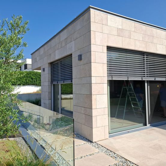 Ein modernes Haus mit Glasfenstern und -türen, einem Glaszaun und einer Person, die im Inneren arbeitet. Das Haus hat eine beige Außenseite und ist von einem Garten mit einem Baum umgeben.