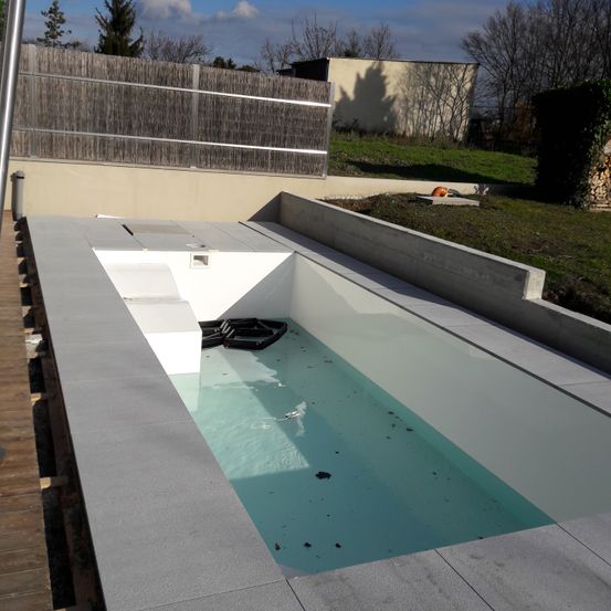 Ein rechteckiger Außenpool, umgeben von Betonwänden, auf einer Holzterrasse. Der Pool hat klares Wasser mit einem schwarzen Objekt, das darin schwimmt.