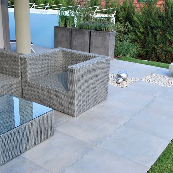 Ein Patio mit Outdoor-Wickelmöbeln, einschließlich eines Stuhls und eines Couchtischs mit Glasplatte, umgeben von großen Betonpflanzgefäßen mit Pflanzen und einem Pool im Hintergrund.
