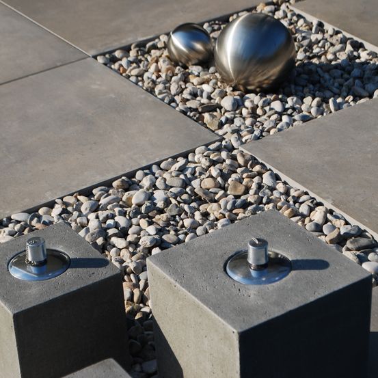 Ein Nahaufnahme einer Betonterrasse mit grauen Fliesen und Kies. Zwei silberne kugelförmige Objekte liegen auf den Kieselsteinen.