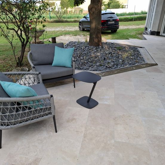 Ein Patio mit Fliesenboden verfügt über ein Set grauer Outdoor-Möbel mit türkisfarbenen Kissen. Ein Baum mit Kies umgibt es, und ein schwarzer SUV ist in der Nähe geparkt.