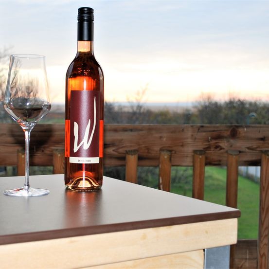 Eine Flasche Wein und ein Weinglas stehen auf einem Holztisch im Freien. Die Umgebung ist eine Terrasse mit einem Holzzaun. Bäume und ein grasbewachsenes Feld sind hinter dem Zaun zu sehen. Der Himmel ist blau mit einigen Wolken.