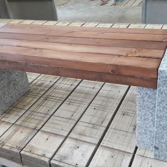 Eine Holzbank wurde auf einem Holzdeck errichtet und wird von Granitblöcken gestützt. Die Bank besteht aus horizontalen Holzbrettern.
