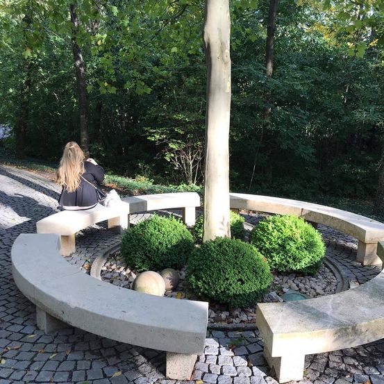 Eine Frau mit langen blonden Haaren sitzt auf einer Bank in einem Park mit einem Baum in der Mitte und Hecken darum herum.