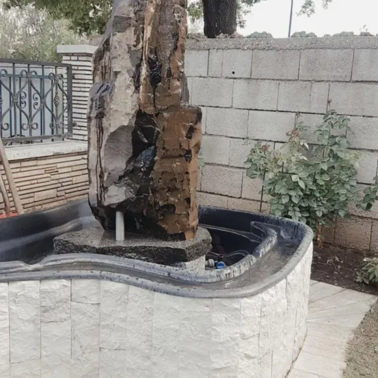 Ein großer, rustikaler Felsbrunnen steht in einem geschwungenen, steingefassten Wasserbecken, umgeben von einer Ziegelmauer und Grünpflanzen.