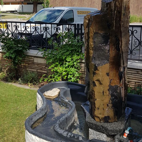 Ein Wasserbrunnen mit einem braunen Steinpfeiler steht in einem Garten. Ein weißer Lieferwagen ist im Hintergrund geparkt.