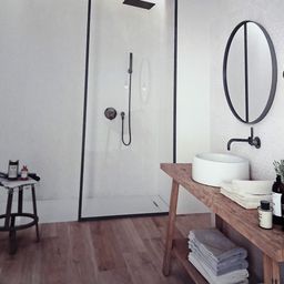 Ein modernes Badezimmer mit einer Glasdusche, einem runden Spiegel, einem Holztisch mit Handtüchern und einem Waschbecken. Der Boden ist aus Holz.