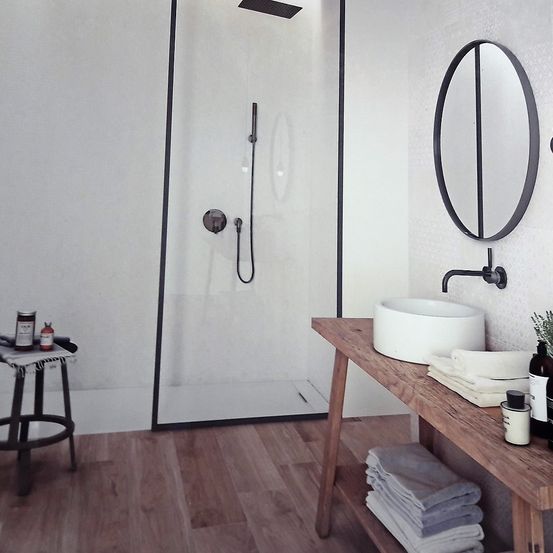 Ein modernes Badezimmer mit einer Glasdusche, einem runden Spiegel, einem Holztisch mit Handtüchern und einem Waschbecken. Der Boden ist aus Holz.