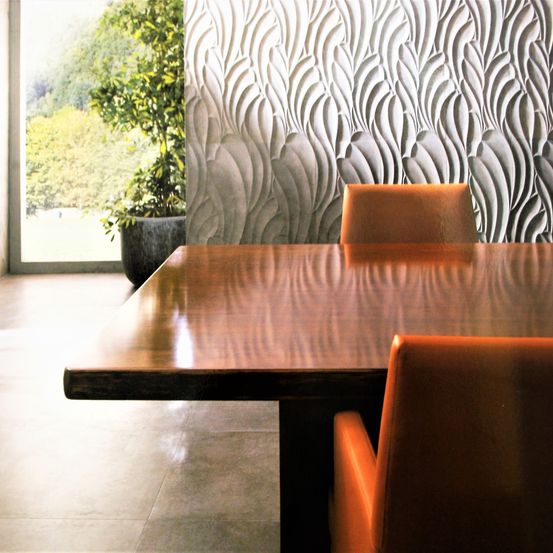 Ein Innenraum mit einem hölzernen Tisch und zwei orangefarbenen Stühlen vor einer strukturierten Wand und einem großen Topf neben einem Glasfenster.