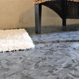 Ein Raum mit grau gefliestem Boden, mit einem kleinen weißen flauschigen Teppich in der Nähe eines Stuhls mit schwarzen Beinen und einer gewebten Sitzfläche.