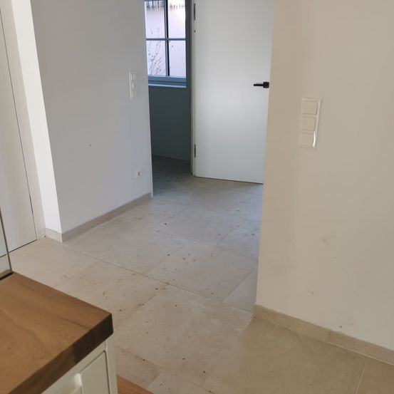 Ein leerer Raum mit weißen Wänden, gefliestem Boden und einer geschlossenen weißen Tür. Ein Fenster ist links sichtbar und ein Schalter ist an der Wand montiert.