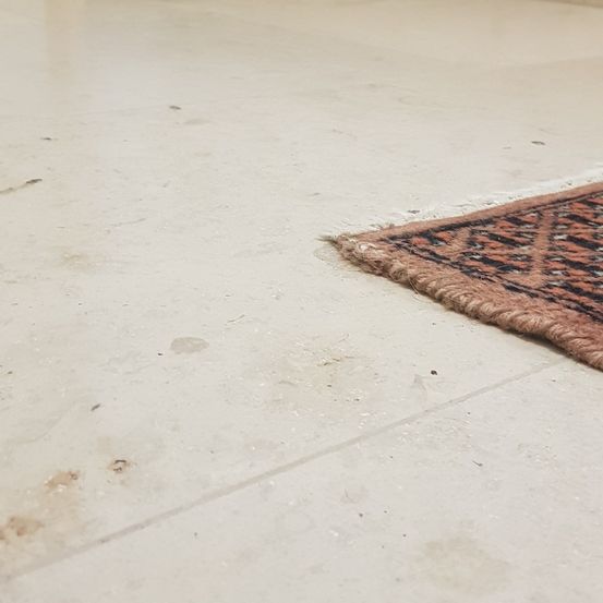 Ein beigefarbener Fliesenboden mit einem kleinen orientalischen Teppich auf der rechten Seite. Der Teppich hat ein geometrisches Muster und der Boden einige Flecken.