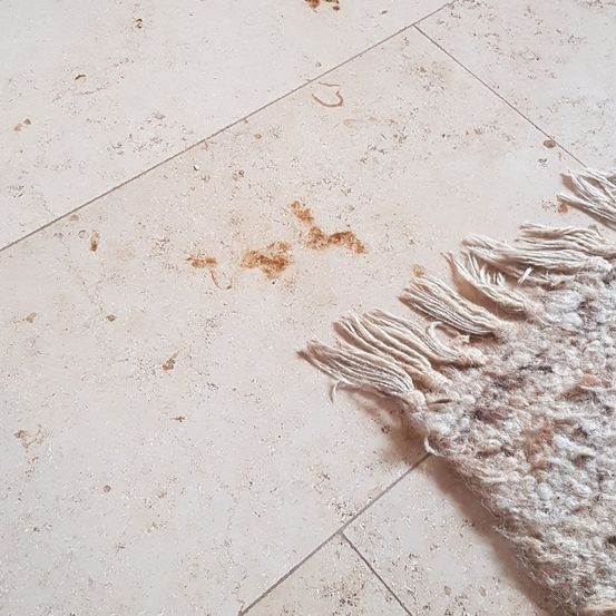 Eine Nahaufnahme eines weißen Fliesenbodens mit braunen Flecken und einem weißen Fransenteppich rechts.