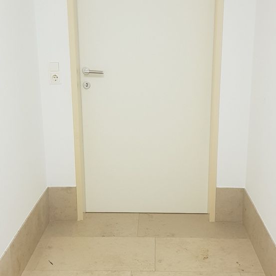 Ein sauberer, leerer Raum mit weißen Wänden, einer weißen Tür und einer Steckdose auf der linken Seite der Tür. Der Boden hat beige Fliesen.