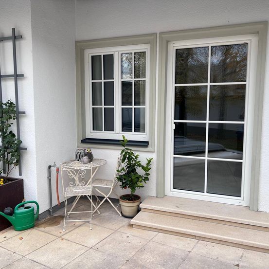 Ein weißer Patio mit einem kleinen Tisch und Stühlen, einem Topfpflanzen und einem Fenster. Das Fenster spiegelt ein Auto und Bäume wider.