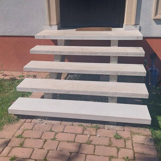 Ein Satz von sechs weißen Betonstufen, die zu einer Tür führen, mit einem Fußabtreter und einem Ziegelweg vor einem Haus.