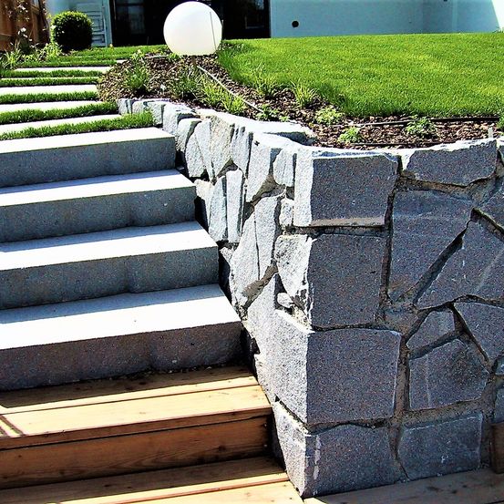 Betonstufen führen zu einer Holzterrasse mit einer Stützmauer aus unregelmäßigen grauen Steinen. Hinter der Mauer befindet sich ein Rasen mit grünem Gras.
