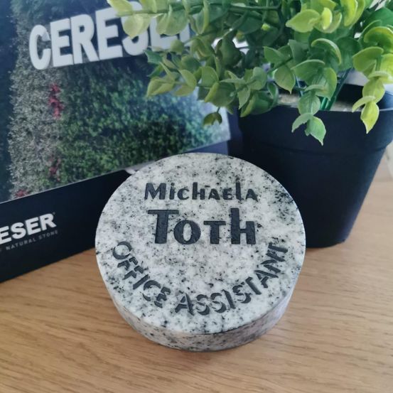 Ein personalisierter Steinstempel auf einer Holzoberfläche mit der Aufschrift 'Michaela Toth Büroassistentin', dahinter eine Pflanze in einem schwarzen Topf. Im Hintergrund befindet sich ein Schild mit dem Wort 'Ceres'.