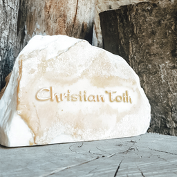 Ein großer, weißer Stein mit dem Namen Christian Toth in Gold eingraviert, auf einer Holzoberfläche platziert. Dahinter befinden sich Baumstämme.
