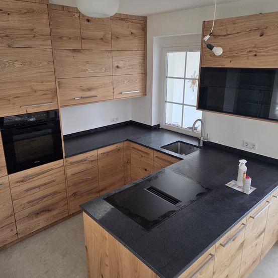 Eine moderne Küche mit Holzschränken, schwarzen Arbeitsplatten, einem schwarzen Herd und einem Waschbecken mit Wasserhahn. Die Küche verfügt auch über ein Fenster und eine Lampe.