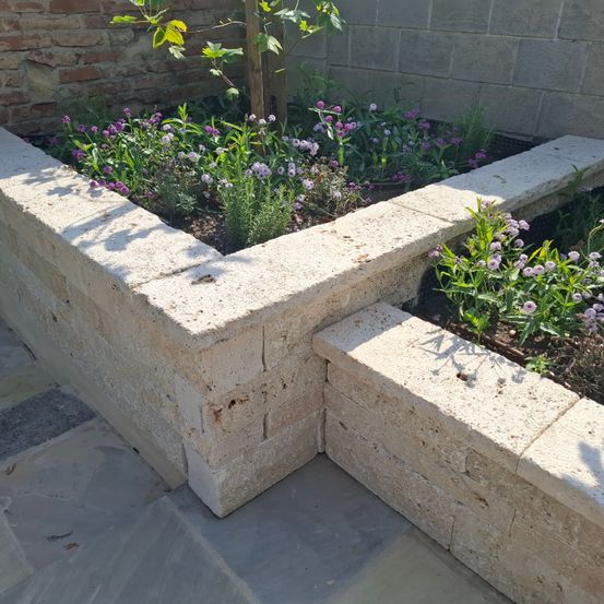 Ein Garten mit Steinmauern, gefüllt mit verschiedenen lila Blumen und Grünpflanzen, umgeben von einer Ziegelmauer.
