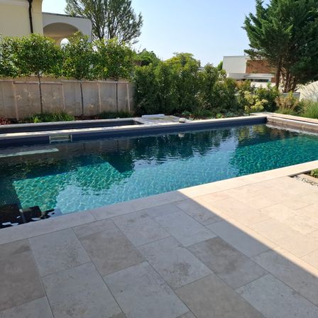 Ein Außenpool mit gefliester Terrasse, umgeben von Bäumen und Pflanzen. Der Pool hat klares blaues Wasser.