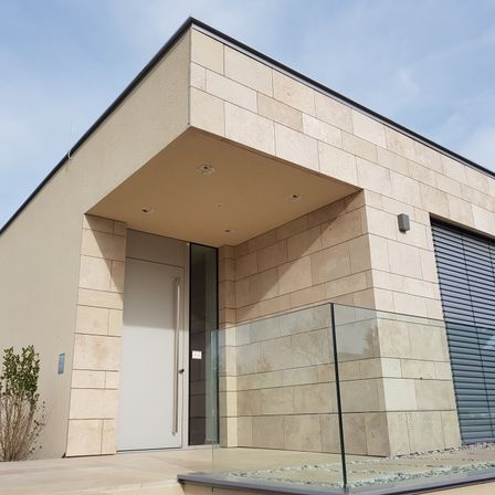 Modernes architektonisches Design mit beigefarbener Ziegelwand und Glaseingang. Eine weiße Tür wird von Glasscheiben flankiert. Eine kleine Pflanze befindet sich links.
