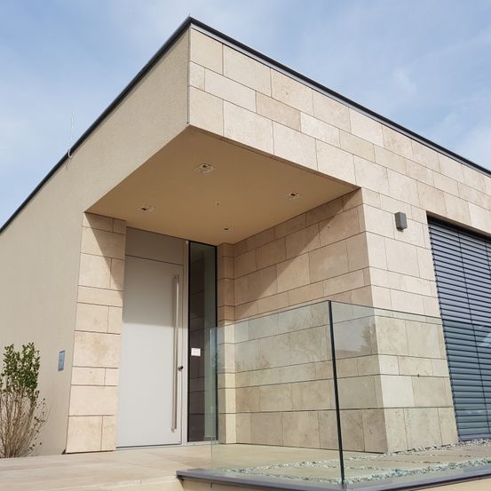 Modernes architektonisches Design mit beigefarbener Ziegelwand und Glaseingang. Eine weiße Tür wird von Glasscheiben flankiert. Eine kleine Pflanze befindet sich links.