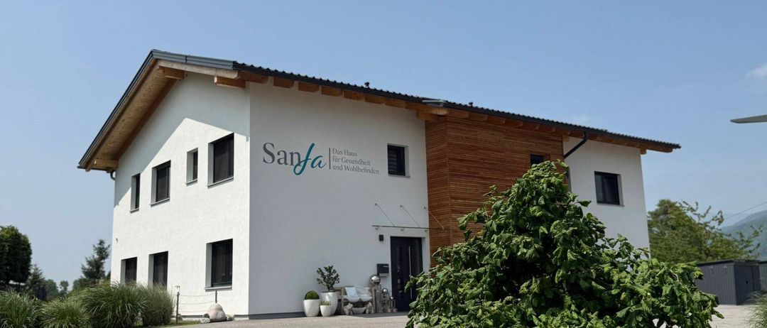 Ein weißes Gebäude namens 'Santa Fe' mit Holzverkleidung steht vor einem blauen Himmel. Es hat mehrere Fenster und eine schwarze Tür. Eine Einfahrt führt zum Gebäude, mit Grünpflanzen und einem Busch im Vordergrund.