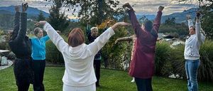 Eine Gruppe von Frauen praktiziert Yoga auf einem Rasen mit Bäumen und Bergen im Hintergrund.