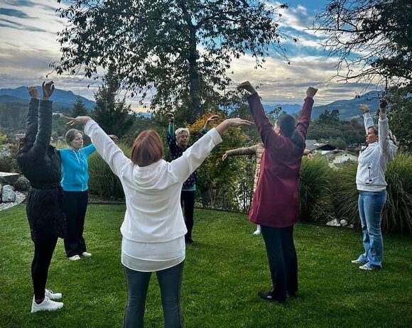 Eine Gruppe von Frauen praktiziert Yoga auf einem Rasen mit Bäumen und Bergen im Hintergrund.